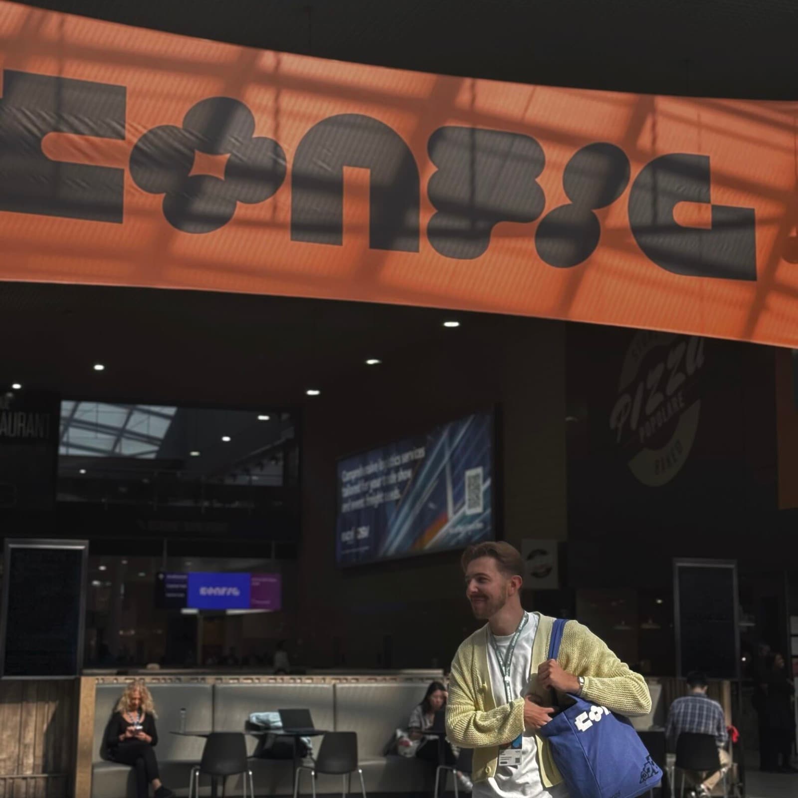 Me under the orange Config banner at the venue entrance, blue Config tote bag on my shoulder