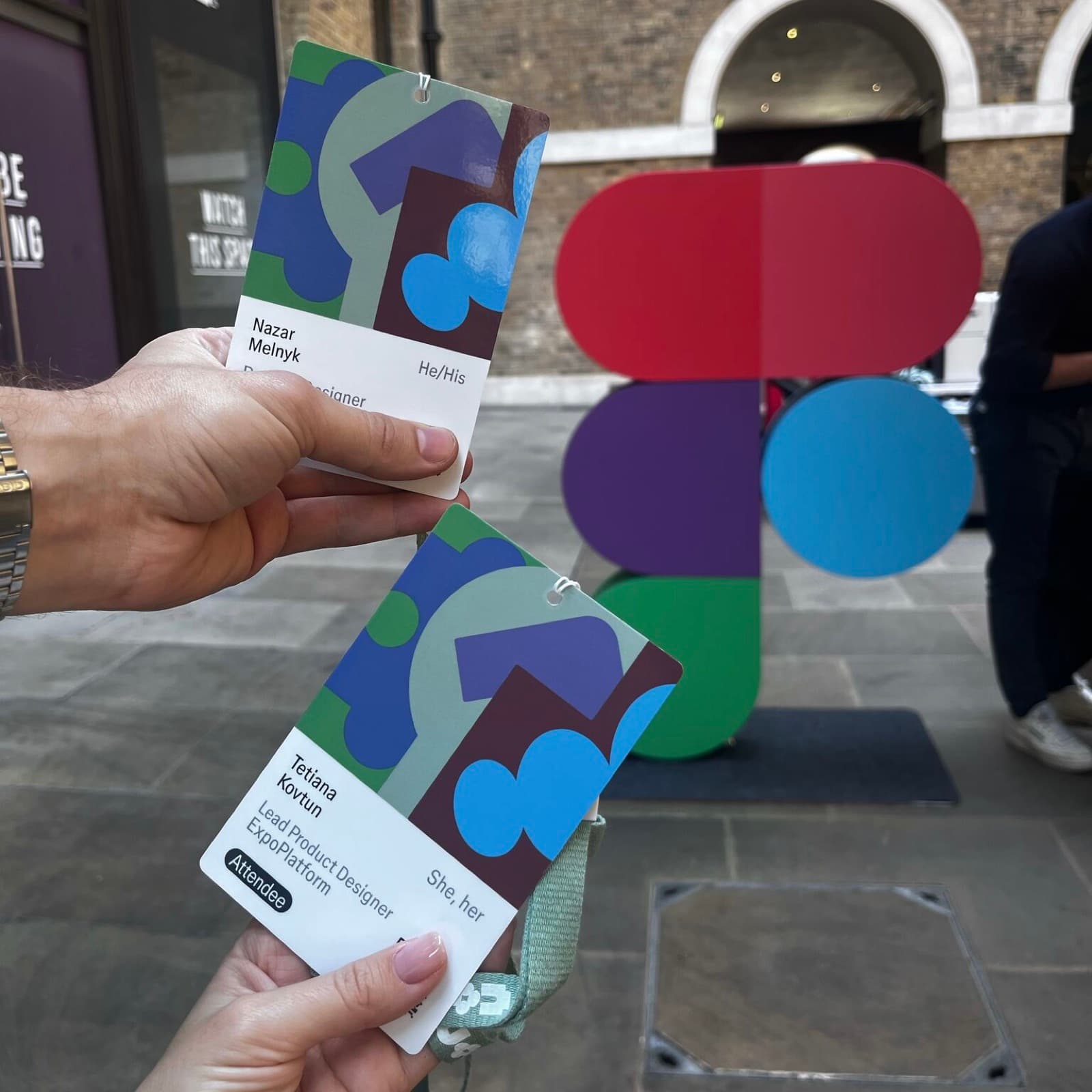 Two attendee badges held up in front of a giant Figma logo sculpture in a London courtyard